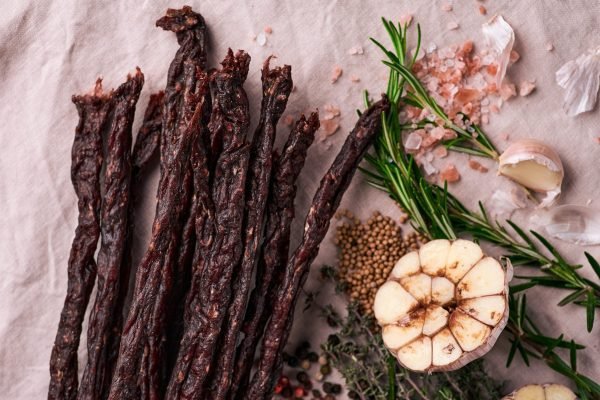 Dried droëwors sticks arranged with fresh garlic, rosemary, coarse salt, and spices on a rustic surface.