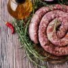 Coiled boerewors sausage seasoned with herbs and spices, displayed on a wooden board with fresh rosemary, chili peppers, and a bottle of oil.