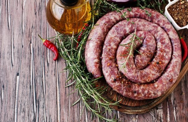 Coiled boerewors sausage seasoned with herbs and spices, displayed on a wooden board with fresh rosemary, chili peppers, and a bottle of oil.