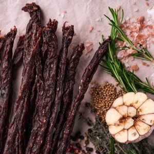 Dried droëwors sticks arranged with fresh garlic, rosemary, coarse salt, and spices on a rustic surface.