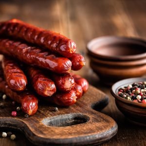 A stack of grilled Kasegrillers on a wooden cutting board with peppercorns in small bowls