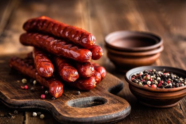 A stack of grilled Kasegrillers on a wooden cutting board with peppercorns in small bowls