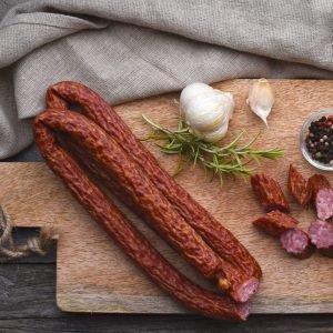 Cabanossi on a wooden cutting board with garlic, peppercorns, and rosemary.