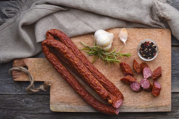 Cabanossi on a wooden cutting board with garlic, peppercorns, and rosemary.