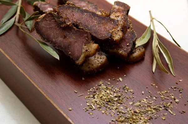 Sliced South African biltong on a wooden board, garnished with dried herbs and olive leaves. A traditional dried meat delicacy known for its rich, spiced flavor.