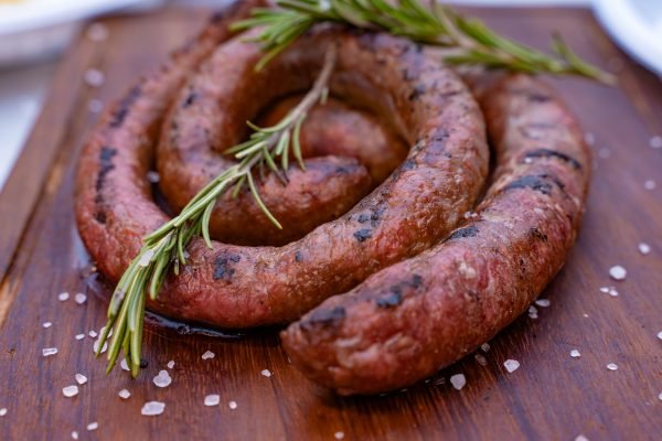 A spiral-shaped grilled Brêkfis Boerewors sausage on a wooden board, garnished with rosemary and sea salt