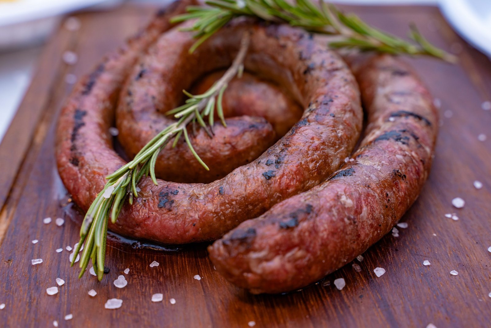 A spiral-shaped grilled Brêkfis Boerewors sausage on a wooden board, garnished with rosemary and sea salt