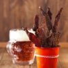 A close-up of a red cup filled with strips of Chutney Biltong Sticks, next to a glass mug of beer on a wooden table