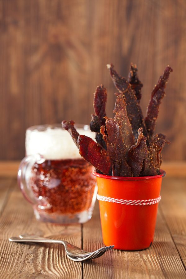 A close-up of a red cup filled with strips of Chutney Biltong Sticks, next to a glass mug of beer on a wooden table