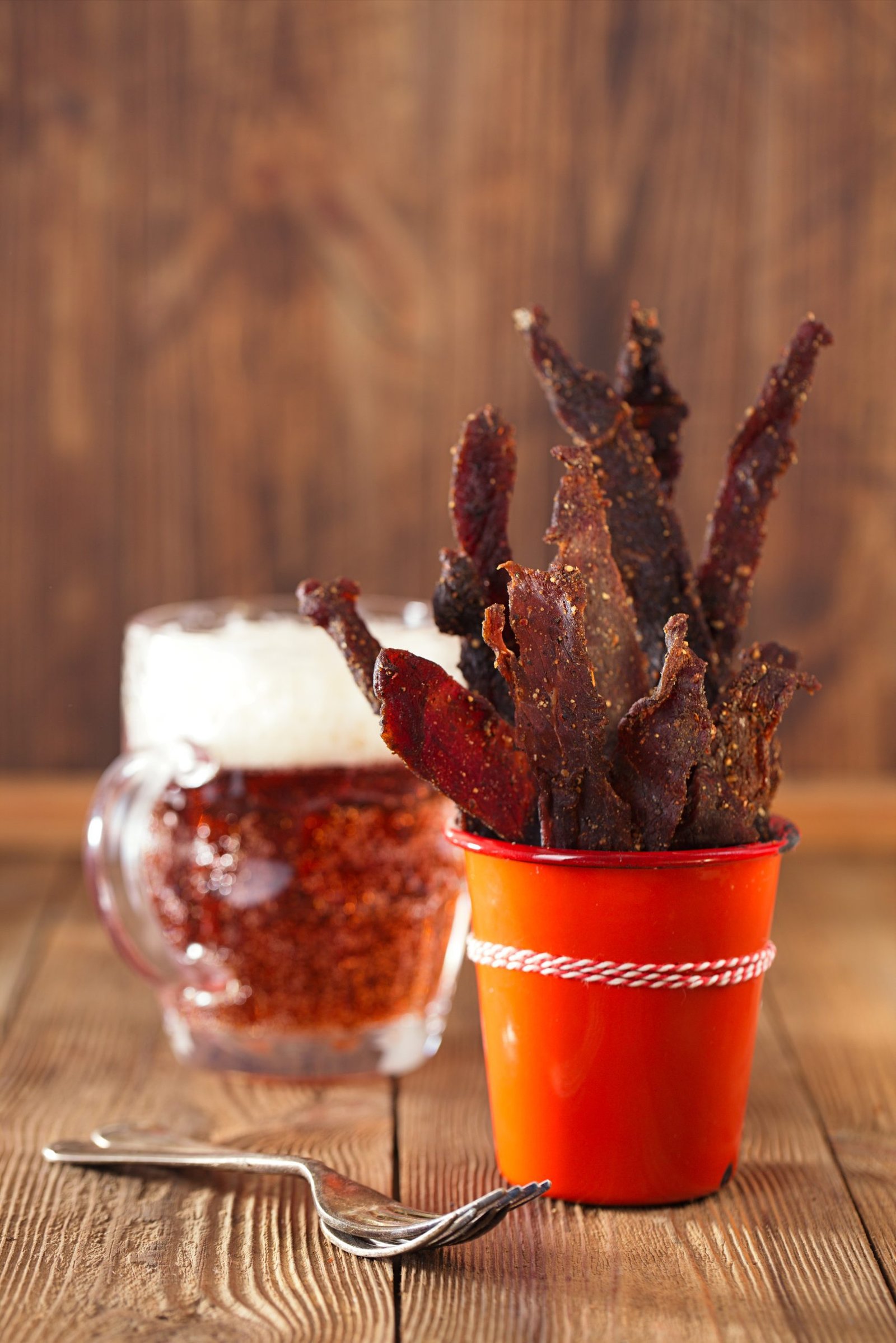 A close-up of a red cup filled with strips of Chutney Biltong Sticks, next to a glass mug of beer on a wooden table